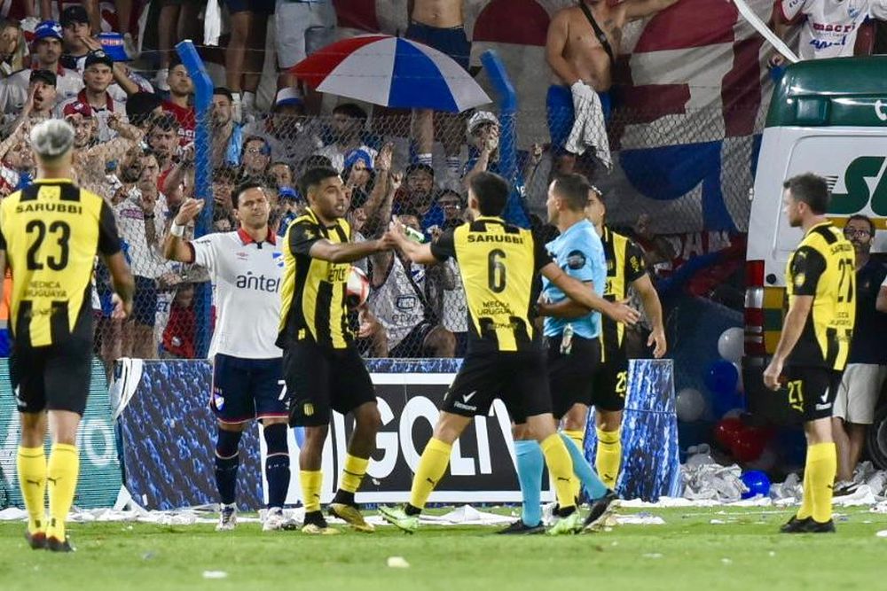 Maximiliano Olivera le entrega al árbitro Ostojich una petaca de vidrio que cayó desde la tribuna cuando el lateral aurinegro intentaba reponer un lateral