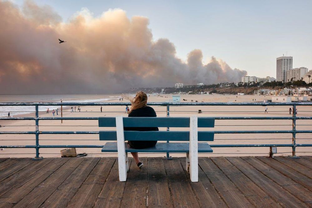Sentada en un banco del muelle de Santa Mónica, una mujer observa la columna de hubo dejada por el incendio en Pacific Palisades.