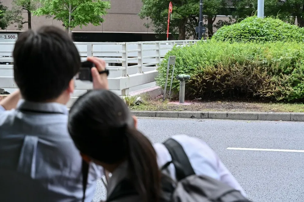 Periodistas nipones se paran frente a un arbusto y pasto quemado, donde el hombre se prendió fuego