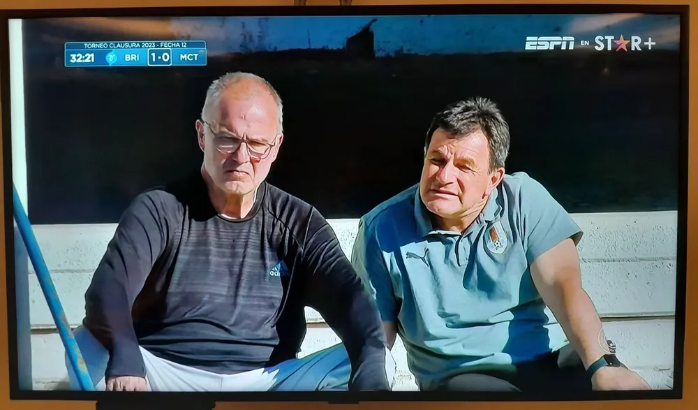Captura de pantalla de la transmisión de Vtv+ en la que se observa a Marcelo Bielsa y a Jorge Giordano en el Parque Artigas de Las Piedras en el partido Boston River vs City Torque